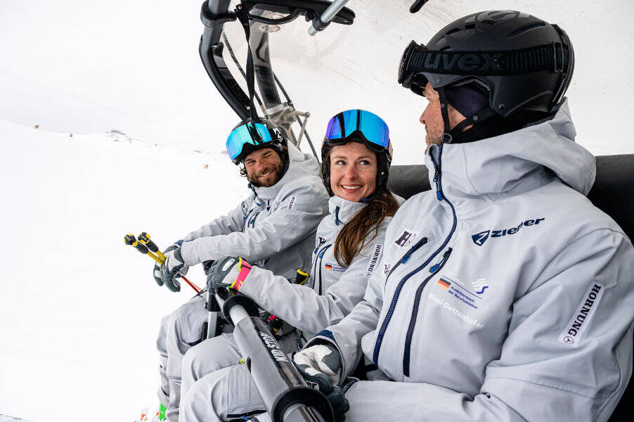 Een vrouw en twee mannen in grijze ski -pakken zitten samen in een stoel. Til de vrouw kijkt naar haar collega met een glimlach