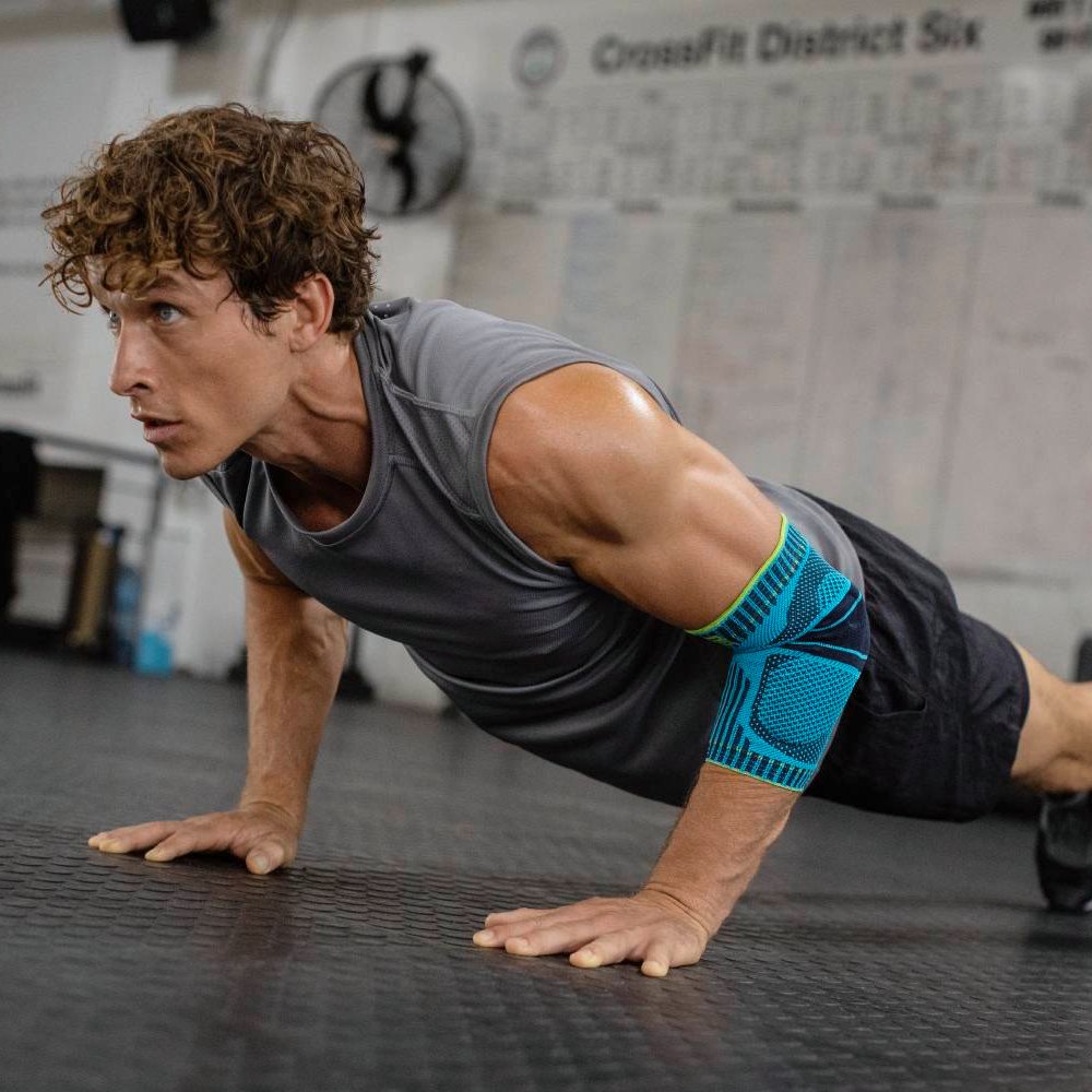 Toon je kracht bij elke push-up! Een sportieve man met een gespierd bovenlichaam voert gefocust push-ups uit in een sportschool. Hij draagt een blauw-zwarte elleboogbandage aan zijn rechterarm voor stabiliteit en ondersteuning tijdens zijn training.