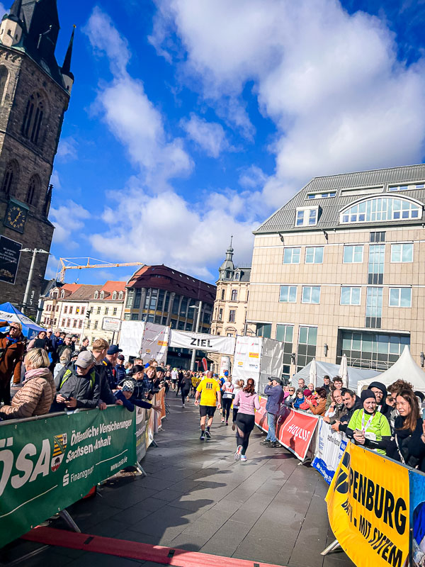 De laatste meters voor de finish bij de halve marathon in Halle, met toeschouwers die de lopers aanmoedigen De laatste meters voor de finish bij de halve marathon in Halle. Lopers passeren de finishlijn onder een blauwe hemel, toegejuicht door het publiek.