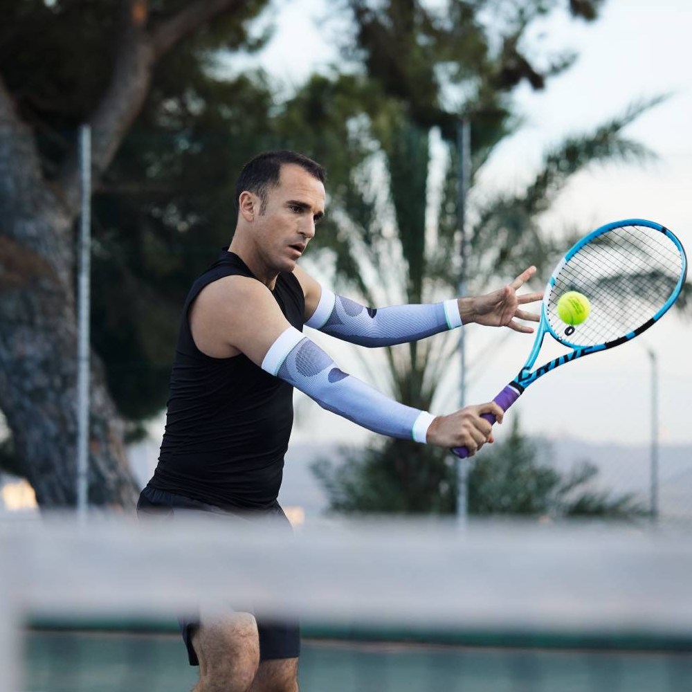 Tennisser met witte compressiemouwen in actie Een tennisser met witte compressiemouwen slaat een forehand. Op de achtergrond zijn bomen en een tennisbaan te zien.