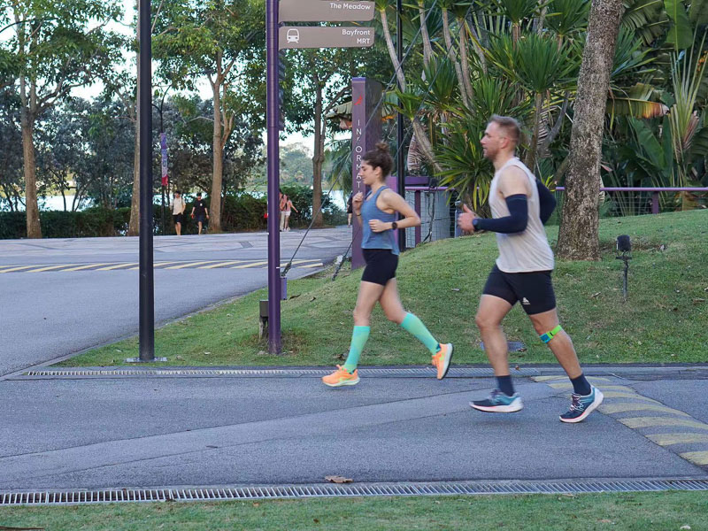 Samen sterk: Hardlooptraining voor de fitnessuitdaging Twee mensen joggen door een stedelijk park op een geasfalteerde weg. Een man in lichte hardloopkleding en een kniebrace rent samen met een jonge vrouw die groene compressiekousen draagt.