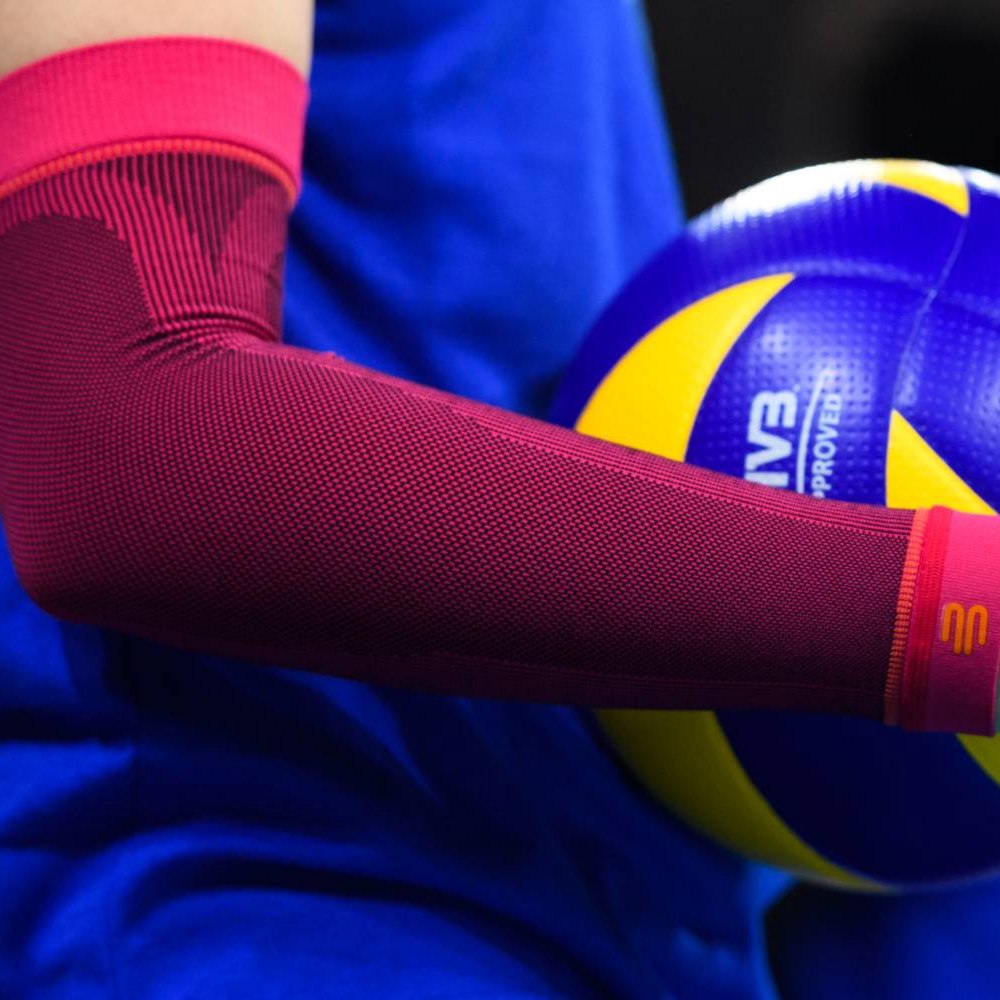 Close-up van een roze volleybal armsleeve op de arm van een speler met een geel-blauwe volleybal op de achtergrond.