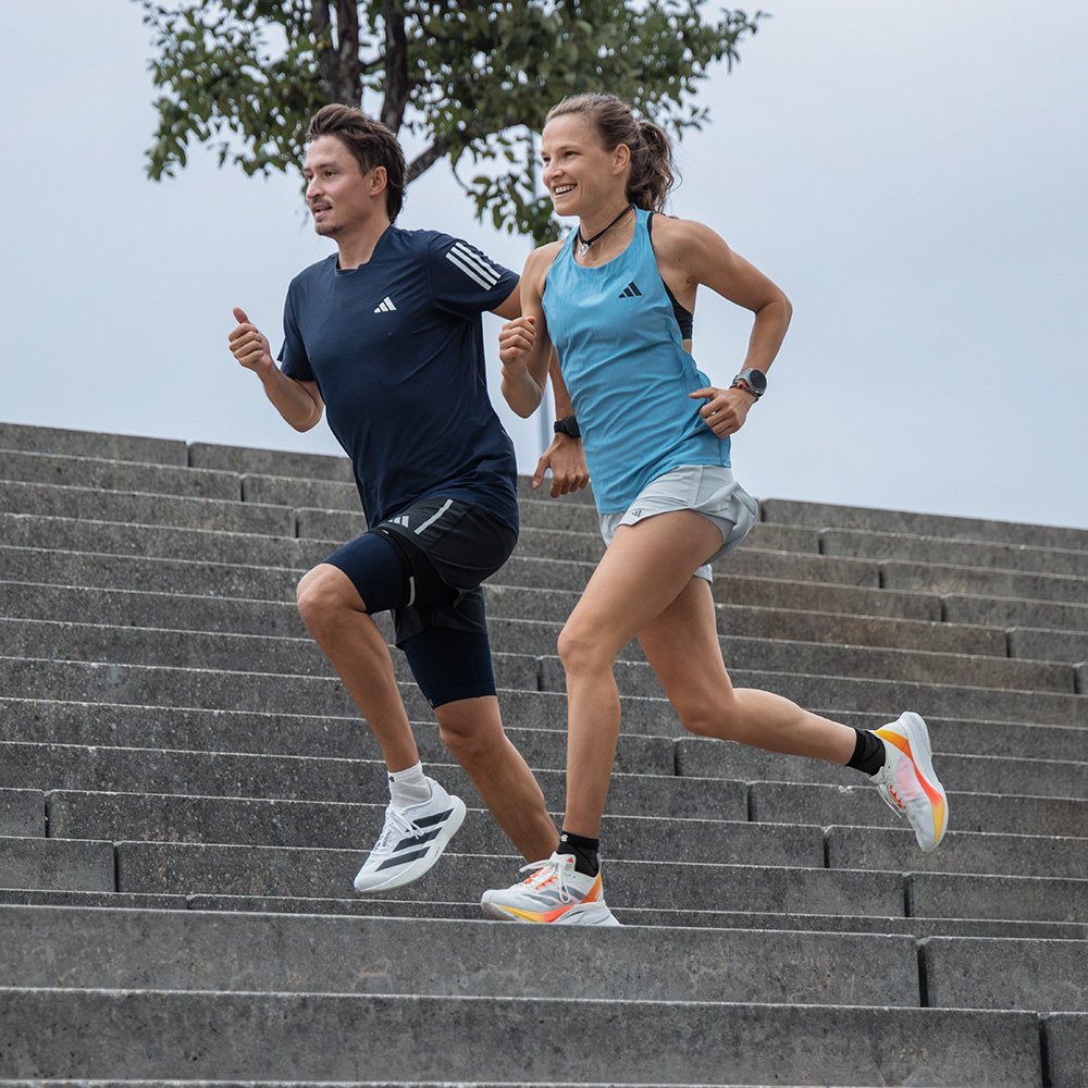 Een sportieve vrouw en een man rennen samen energiek een brede stenen trap af. Beiden dragen sportkleding en schoenen van adidas.