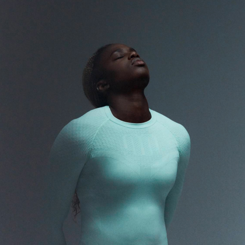 Compressieshirt in actie – studio-opname Een persoon staat in een oranje verlichte studio met de handen in de zij, gekleed in een strak blauw compressieshirt dat de romp en armen volledig bedekt.