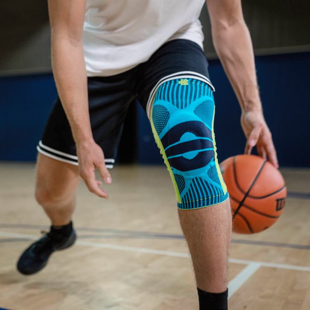Basketbaltraining met turquoise kniebandage Een basketbalspeler met een turquoise kniebandage op zijn rechterbeen dribbelt de bal in een sporthal; de nadruk ligt op het product en zijn houding.