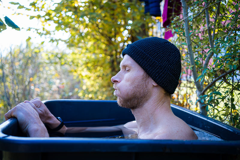 Man tijdens ijsbaden in een buitenbad. Een man met een donkere gebreide muts zit tot aan zijn schouders in een ijsbad in de buitenlucht. Hij is omringd door herfstige vegetatie en zijn gezichtsuitdrukking toont diepe ademhaling om zich aan de kou aan te passen.