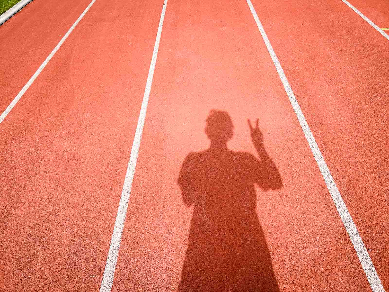Schaduw op de Renbaan – Succes en Doelbereiking na Hardlooptraining. De schaduw van een loper op een rode renbaan die het victory-teken met zijn hand toont. De scène symboliseert succes en het behalen van doelen tijdens hardlooptraining.