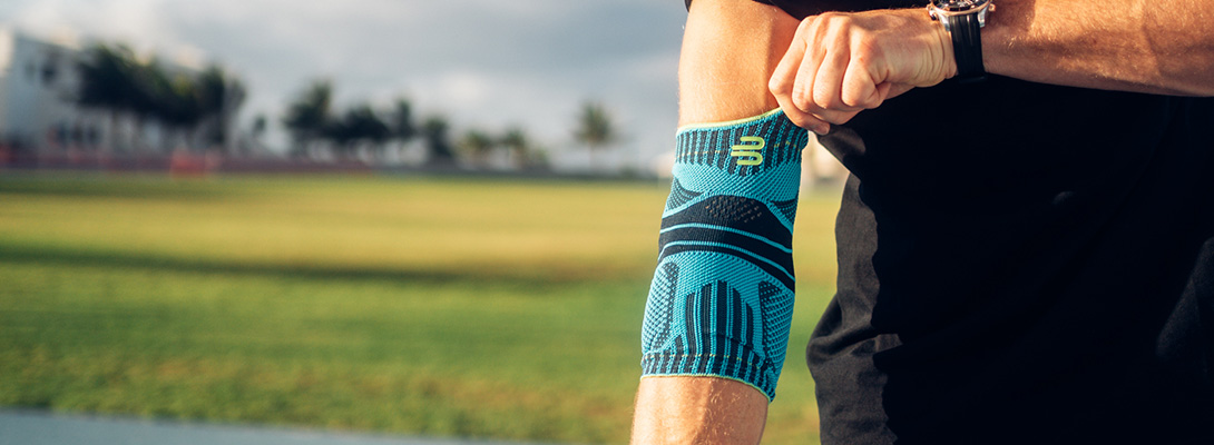 Maak je klaar voor je grote worp! Close-up van een sporter die zich concentreert terwijl hij een blauw-zwarte elleboogbandage om zijn rechterarm doet. Op de achtergrond is een zonnig sportveld te zien.
