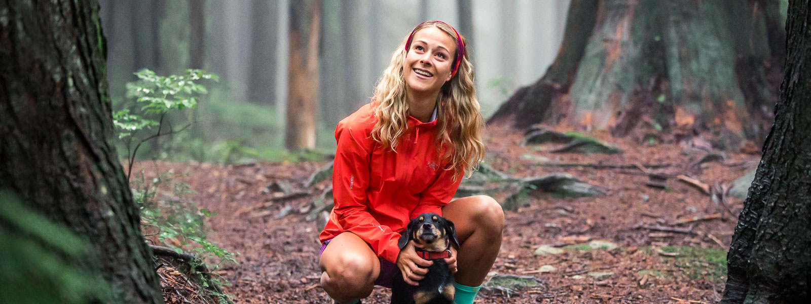 Vrouw met een rood lopende jas en hoofdband houdt haar kleine hond op een bospad en glimlacht