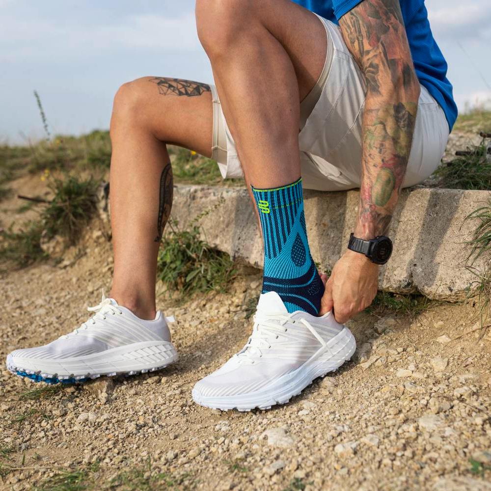 Voorbereiding op de run: veters strak, focus aan! Een man zit op een betonnen rand in een natuurlijke omgeving met een grindpad en gras. Hij draagt witte hardloopschoenen en blauwe sportsokken met een opvallend patroon. Zijn handen trekken de schoen over de sok terwijl zijn getatoeëerde arm zichtbaar is.