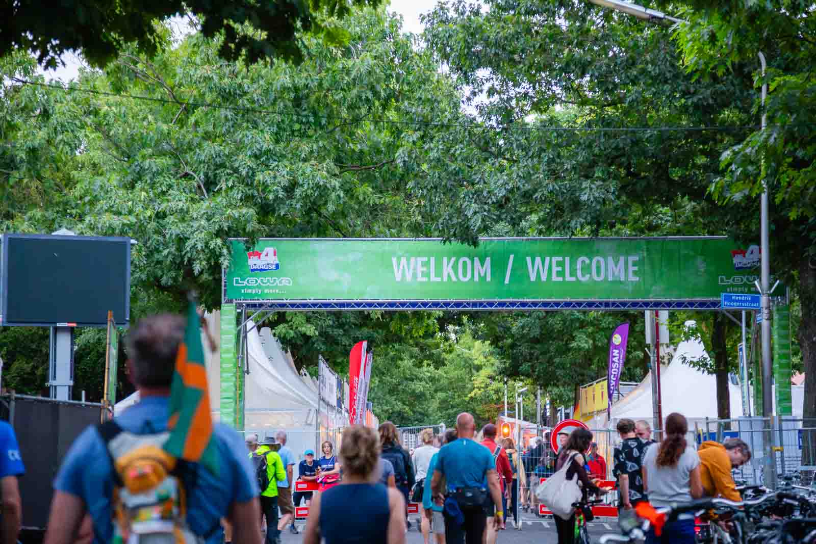 Toegang tot de 4Daagse in Nijmegen met een groen spandoek „Welkom / Welcome“ en wandelaars op weg naar het startgebied.