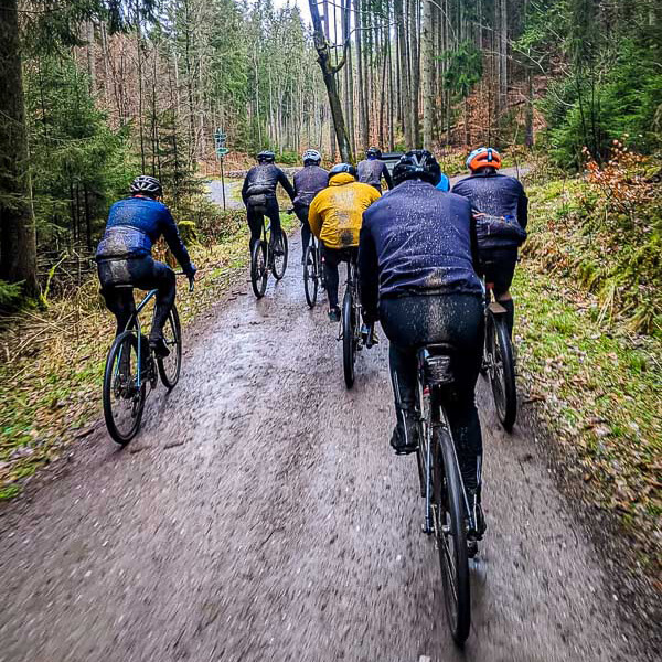 Modder, zweet en teamgeest – als de fitnesschallenge het terrein in gaat! Een groep fietsers rijdt over een modderig bospad, omgeven door hoge naaldbomen. De fietsers zijn van achteren te zien en dragen weerbestendige kleding en helmen.