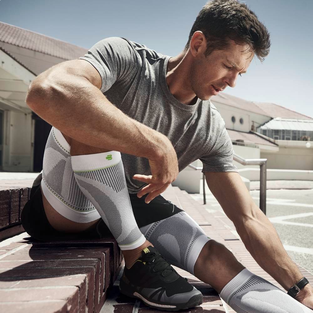 Een gespierde man zit op een trap buiten in de zon, draagt witte compressiekousen en lijkt te herstellen van een intensieve run. Hij haalt diep adem en bereidt zich mentaal voor op de volgende uitdaging.