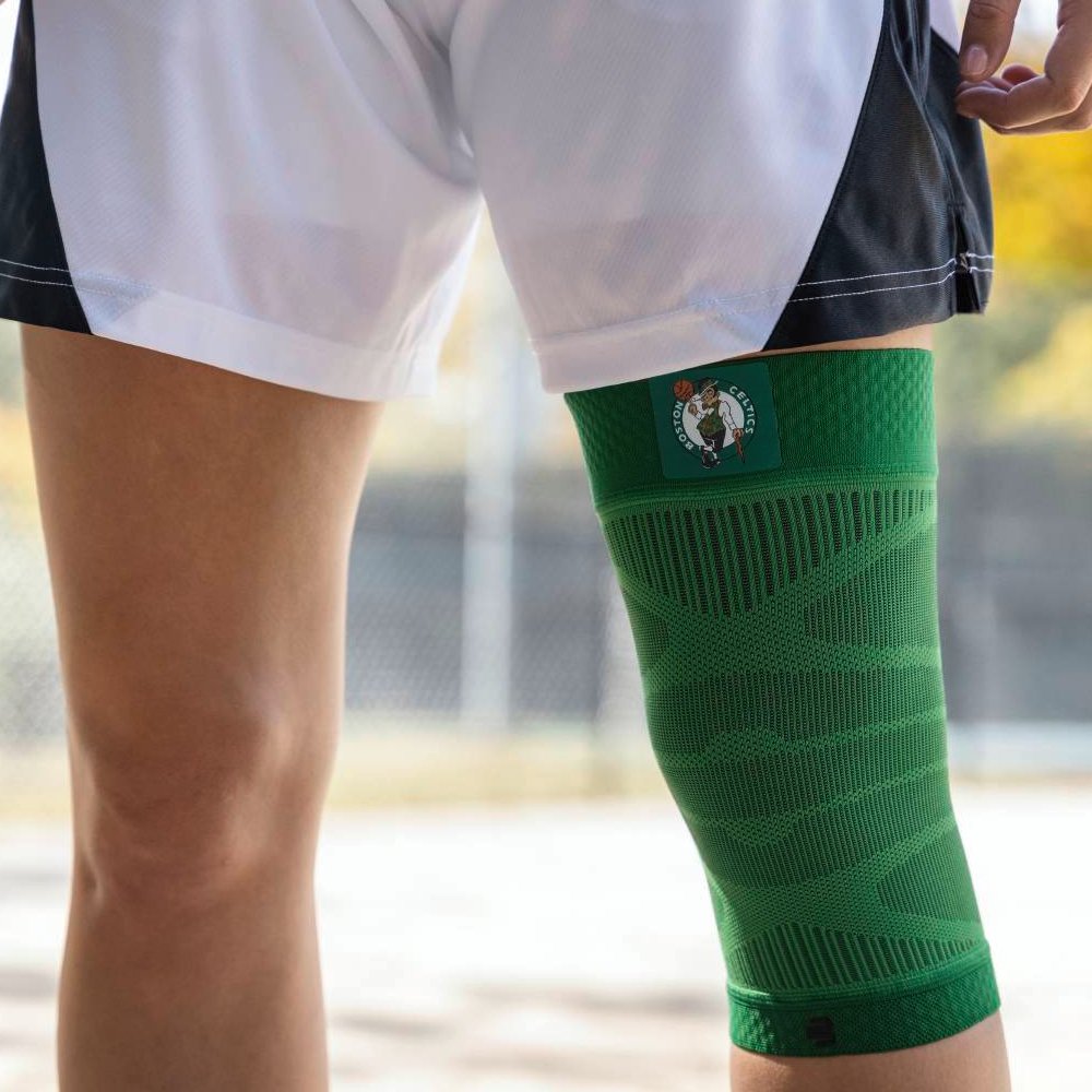 Close-up van een speler met een groene Boston Celtics-kniebrace op een basketbalveld. De brace toont het teamlogo en past ergonomisch om de knie.