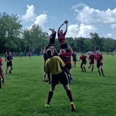 Abheben für den Sieg! Ein Sprung, der das Spiel entscheidet. Ein Rugby-Spiel in vollem Gange auf einem grünen Rasenfeld. Ein Spieler in rot-schwarzem Trikot wird von Teamkollegen hochgehoben, um den Ball zu fangen, während der Schiedsrichter in gelber Kleidung die Szene beobachtet.