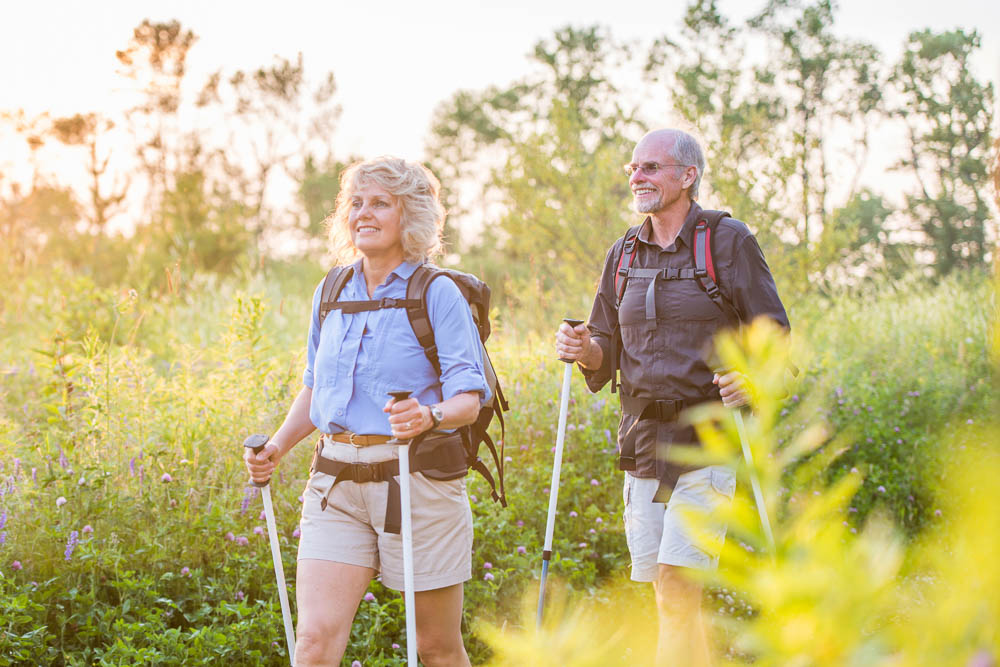 Actief wandelen in zomerlicht Een ouder stel wandelt met trekkingstokken door een bloeiende zomerse weide in het warme avondlicht. Beiden dragen lichte wandelkleding en rugzakken en zien er tevreden uit. De scène straalt rust, vitaliteit en wandelplezier uit.