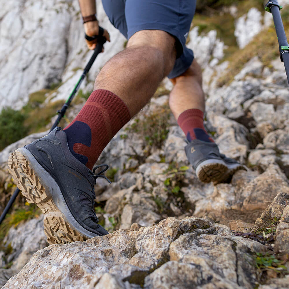 Close-up van een wandelaar met Merino Outdoor Socks in granaatkleur terwijl hij een rotsachtig pad beklimt. De sokken bieden ondersteuning en comfort op uitdagend terrein.