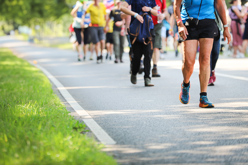 Wandelgroep tijdens de 4Daagse Een groep wandelaars loopt over een afgesloten weg in een groene omgeving op een zonnige dag. Op de voorgrond is een man in blauwe sportkleding en hardloopschoenen te zien.