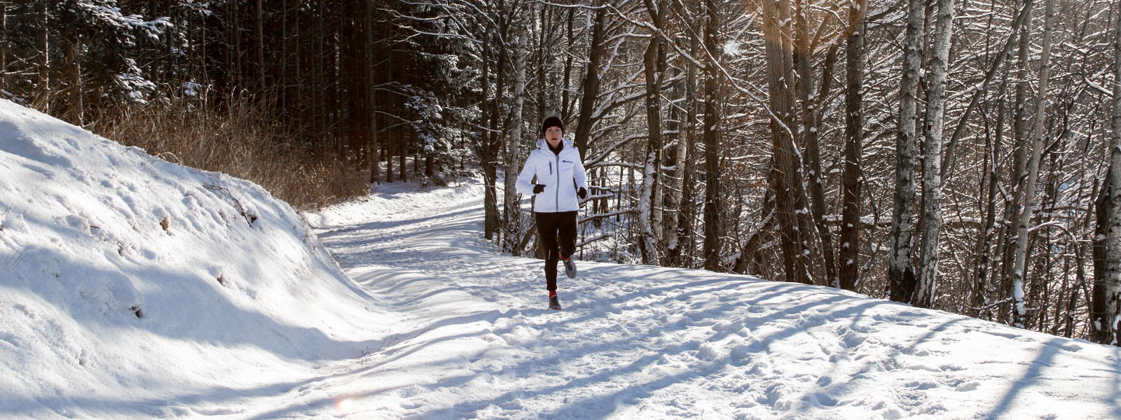 Vrouw in een wit hardloopjasje joggt op een met sneeuw bedekt pad tussen bomen en kijkt naar de camera