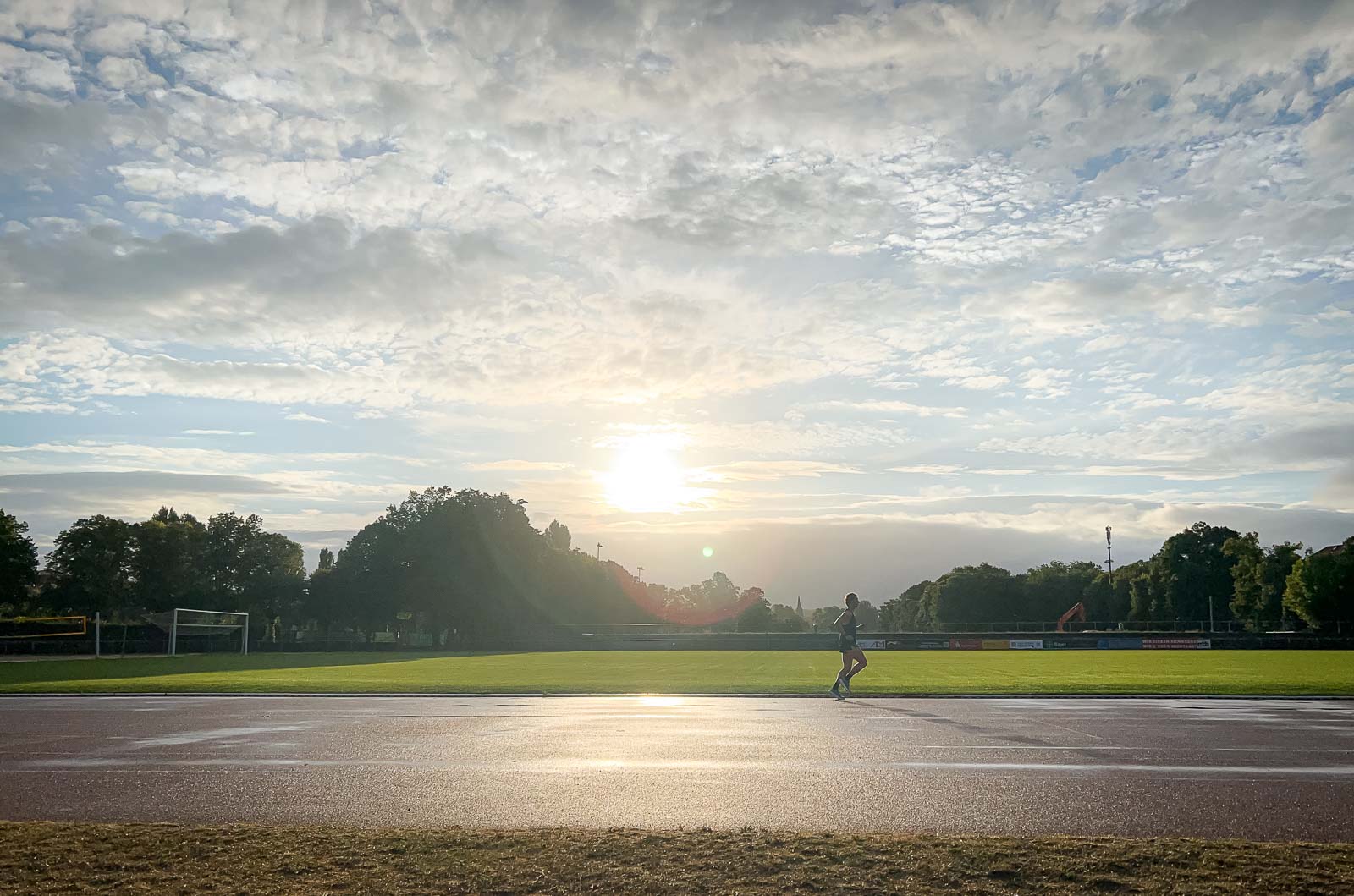 Loper op de Renbaan – Zonsopgangstraining voor Uithoudingsvermogen en Snelheid. Een loper op een natte renbaan bij zonsopgang. De lucht is bewolkt, maar de zon breekt door terwijl de loper zijn ronde afmaakt.