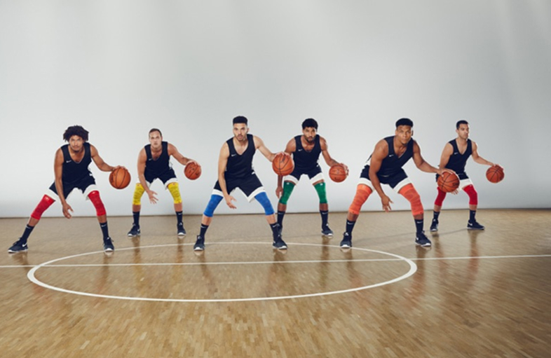 Zes basketballers staan op een indoorveld in een rij en zijn gebukt in een voorbereidingshouding. Ze dragen zwarte shirts, witte shorts en compressiemouwen in verschillende kleuren om hun knieën. 
