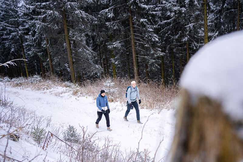 Winterwandelen – Waar elke stap een krakend avontuur is. wee vrouwen wandelen door een besneeuwd boslandschap. Ze dragen warme winterkleding en gebruiken wandelstokken terwijl ze een pad volgen. Op de achtergrond zijn met sneeuw bedekte bomen en een dicht bos te zien.