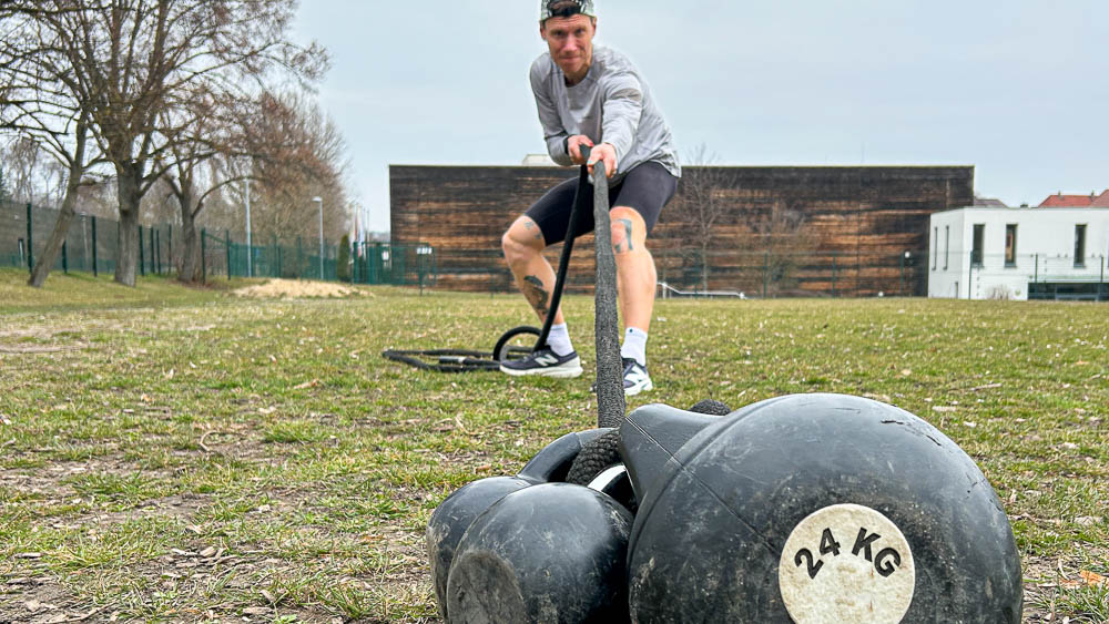 Atleet sleept tijdens HYROX-training buiten twee kettlebells met een band over het gras – functionele krachttraining zonder sportschool.