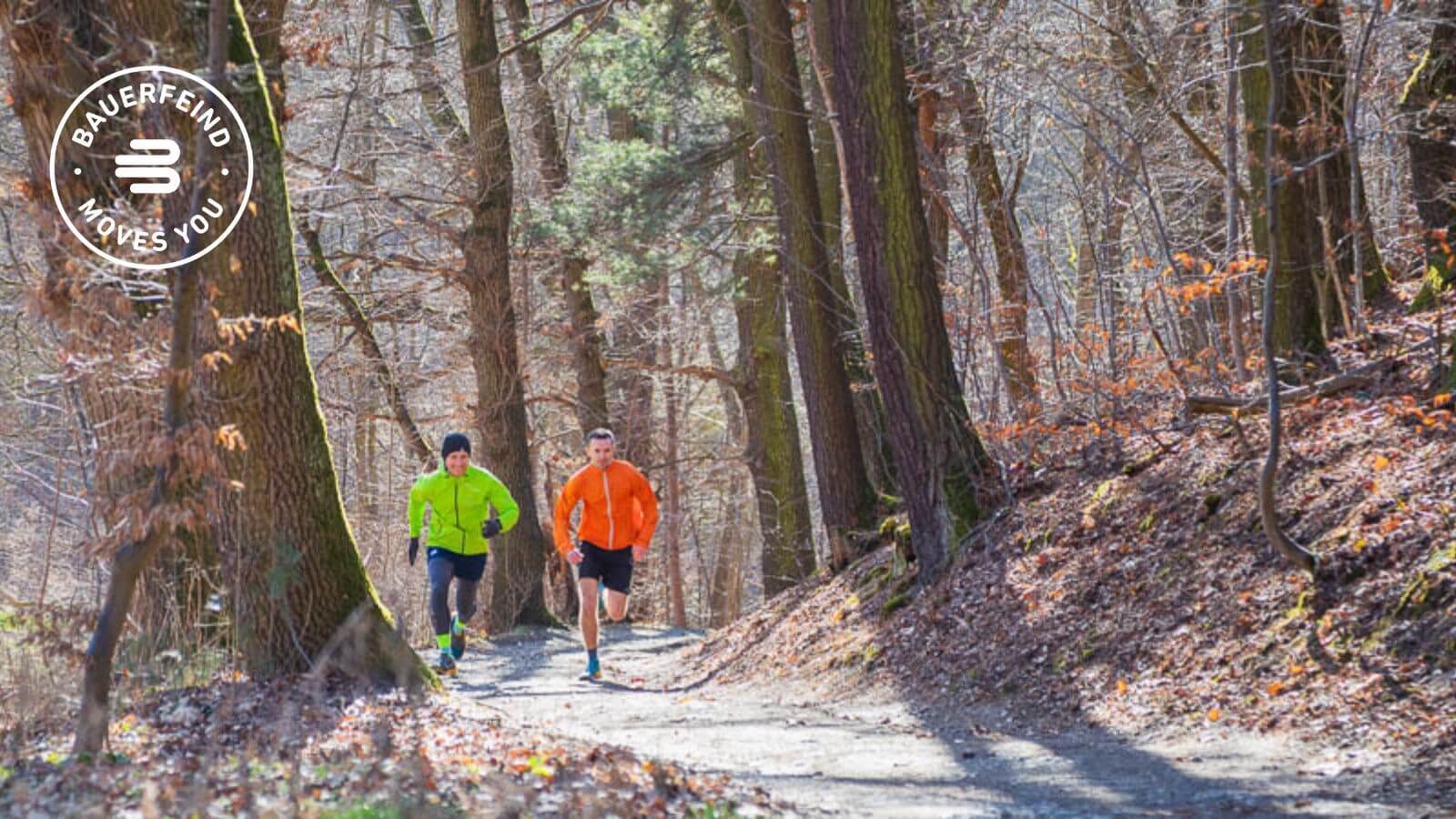 Pure motivatie - op snelheid door het bos Twee hardlopers in felgekleurde jassen (groen en oranje) joggen over een zonnig bospad, omringd door hoge bomen en droog bladerdek. Beiden dragen opvallende sokken, wat op functionele sportkleding kan wijzen.