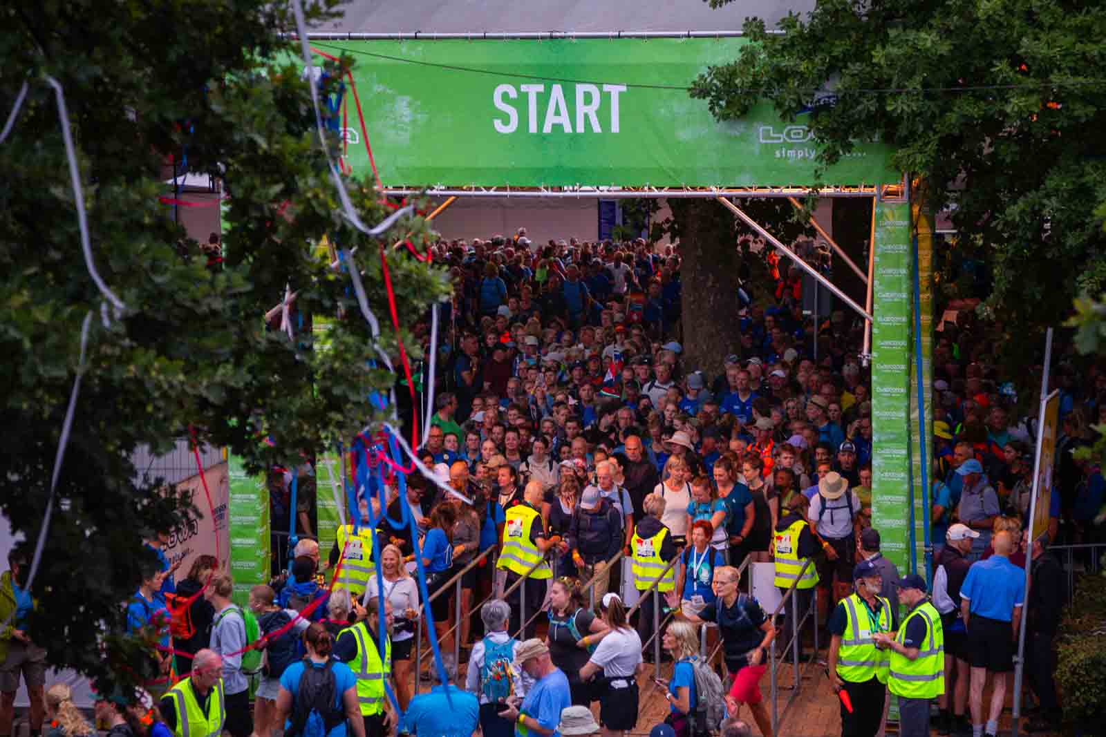 Deelnemers van de 4Daagse in Nijmegen verzamelen zich bij de drukke start onder een groot groen spandoek met de tekst „START“.