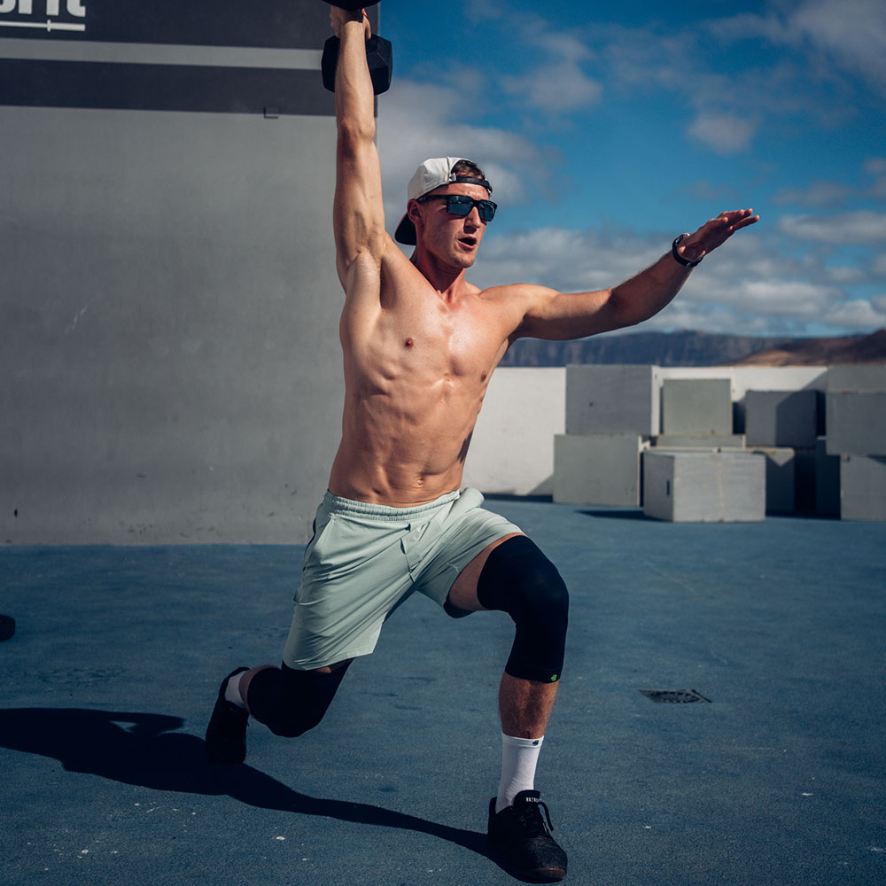 Maximale stabiliteit – zelfs bij intensieve trainingen! Een fitte man doet een eenbenige lunge met een halter boven zijn hoofd, terwijl hij een zwarte kniesteun draagt.