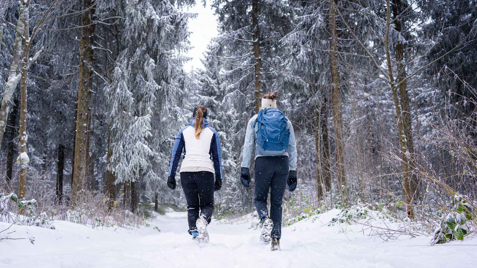 Wintermagie: Volg het pad naar het witte avontuur Twee vrouwen lopen door een dik besneeuwd bos, gezien van achteren. Ze dragen winterjassen, wandelschoenen en handschoenen. Een van hen draagt een blauwe rugzak.