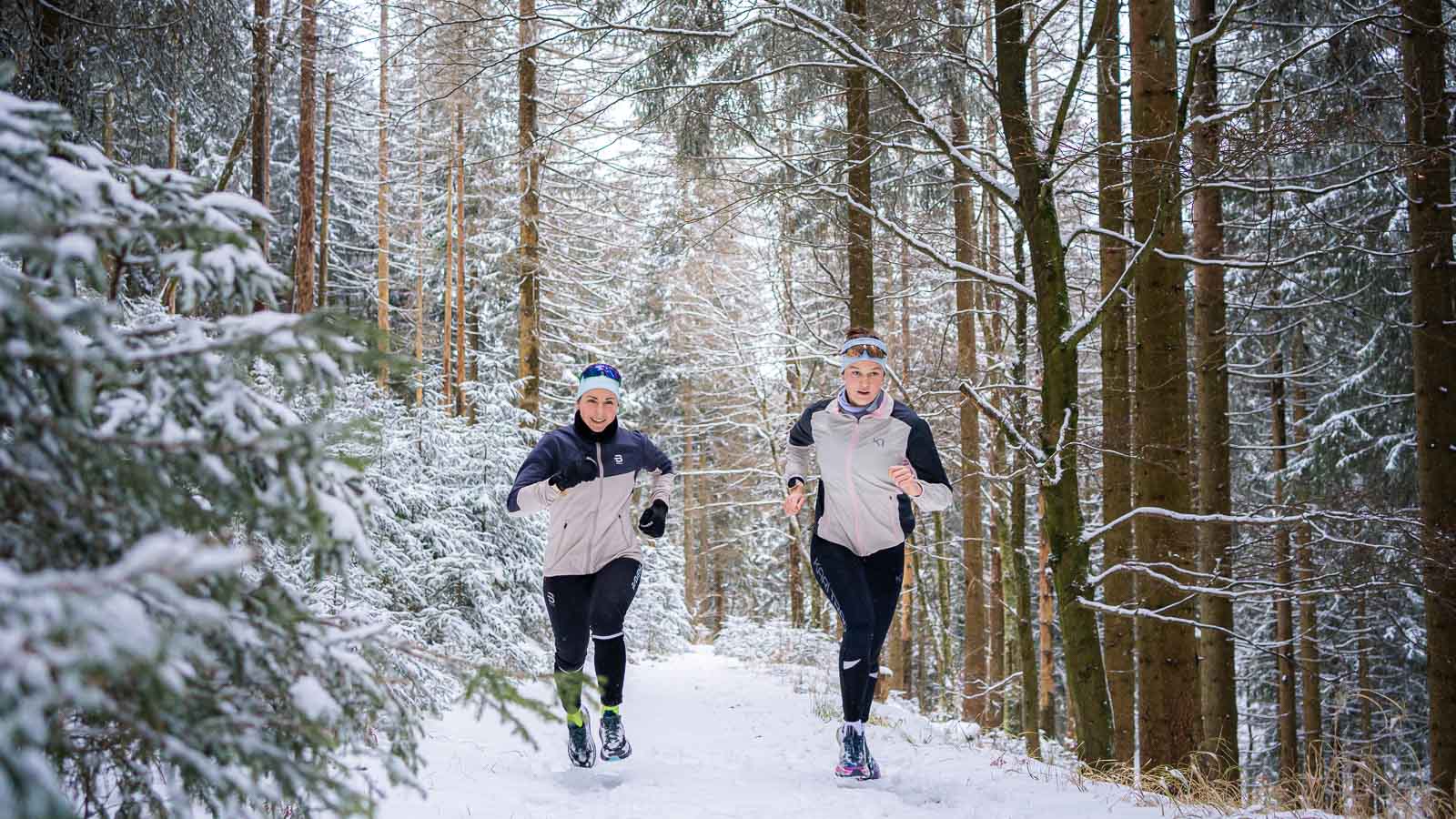 Hardlopen door een winters dennenlandschap Twee hardlopers joggen op een met sneeuw bedekt bospad. De besneeuwde sparren vormen een dicht, winters decor. Beiden dragen warme jassen en hoofdbanden voor een winterse hardlooptocht.
