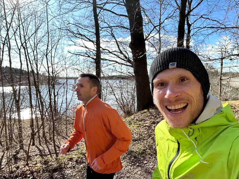 ennend door de natuur Twee mannen rennen op een zonnig bospad langs een meer. De een draagt een neongroene jas en een muts, de ander een oranje jas.