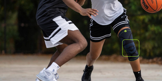 Twee basketballers in actie op een buitenveld. De voorste speler met zwarte kniebrace dribbelt en wordt verdedigd door een tegenstander. De foto laat de dynamiek en het spelritme duidelijk zien.