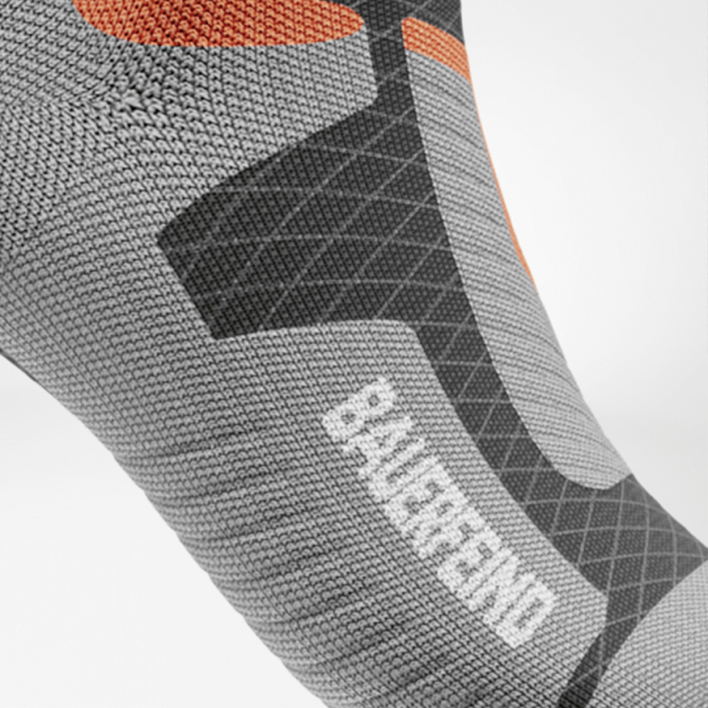 Close-up van het voetgedeelte van de sok met grijze, oranje en zwarte verstevigingen en het BAUERFEIND-logo op de wreef.