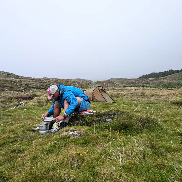 Fitness-uitdaging: Wanneer de natuur je keuken wordt en elke maaltijd een avontuur is. Een persoon in een blauwe jas zit op een weide in heuvelachtig terrein voor een kleine tent en bereidt een maaltijd op een campingkooktoestel. De lucht is bewolkt en het landschap oogt ruw en ongerept.