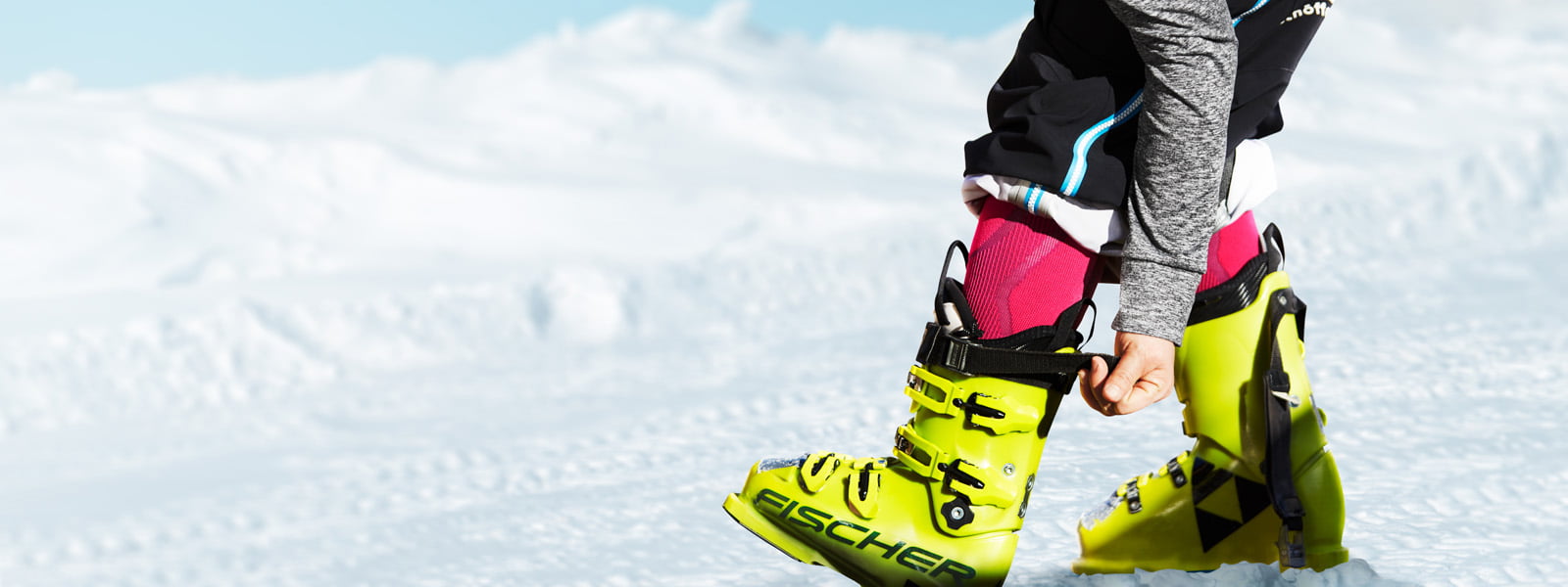 Skiërs met roze ski -sokken en grijze mouwen trekken de riem op zijn gele ski -laars	 sneeuw op de achtergrond