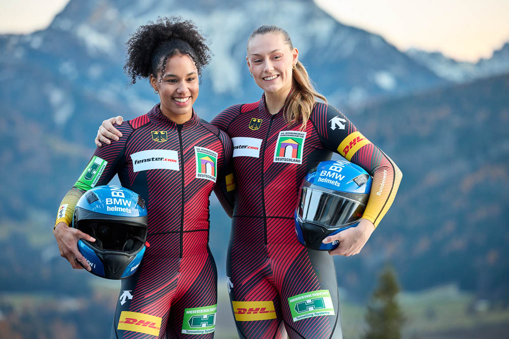 Twee professionele sporters in Duitse nationale bobslee-pakken staan naast elkaar tegen een bergachtig Alpenlandschap en houden hun helmen vast. Ze stralen focus en teamspirit uit.