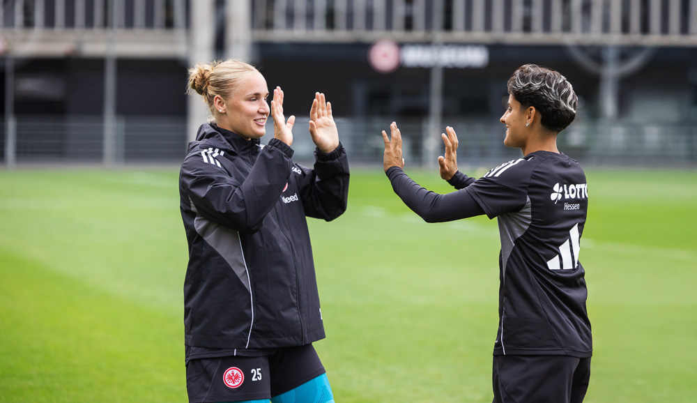 Twee voetbalsters van Eintracht Frankfurt geven elkaar een high five op het veld, gekleed in trainingskleding. Ze dragen beide compressiebandages van Bauerfeind Sports, een teken van professioneel gebruik.