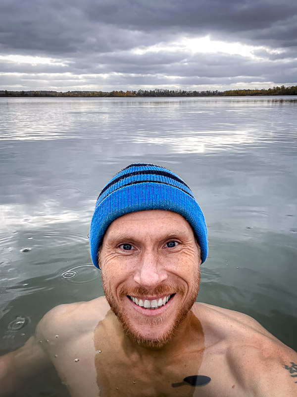 Lachen met kippenvel – de ultieme duik in koud water! Een man met een blauwe muts lacht naar de camera terwijl hij in een koude meer staat. De lucht is bewolkt en het rustige water weerspiegelt de grijze wolken.