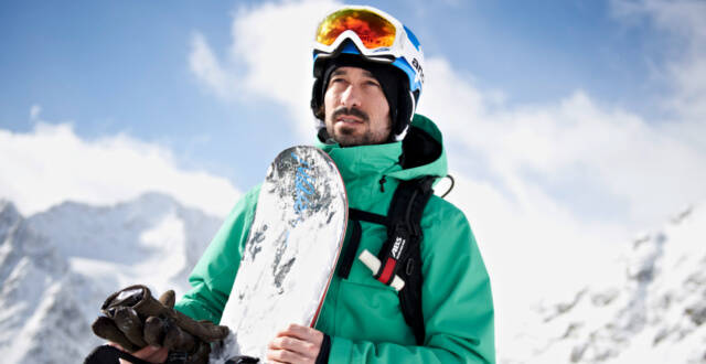 Snowboarder met een groen jasje met bord in de hand voor een bergachtige achtergrond