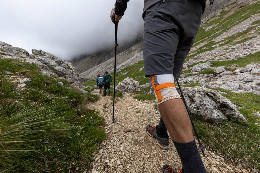 Wandelen in uitdagend terrein - Uitgerust met trekkingstokken en kniebrace! Een wandelaar met trekkingstokken en een kniebrace loopt op een rotsachtig pad door een bergachtig landschap. Andere wandelaars en wolken boven de bergen zijn op de achtergrond te zien.
