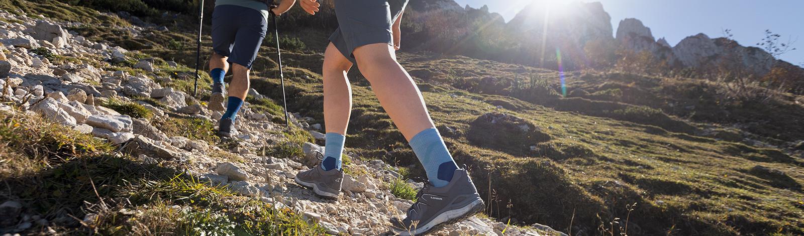 Veiliger meer voorkomt in het ruwe terrein: onze Outdoor Performance Mid Cut Socks Sluit bekleding van een wandelaar in wandelschoenen en met groene middelgrote wandelsokken die een steenachtige pad op de achtergrond hebben, haar wandelpartner