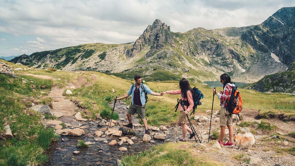 Samen door ruig berglandschap Drie wandelaars, twee vrouwen en een man, steken samen een bergbeek over in een alpenlandschap met rotsachtige toppen op de achtergrond. De man helpt een vrouw – de nadruk ligt op teamwork en natuurbeleving.