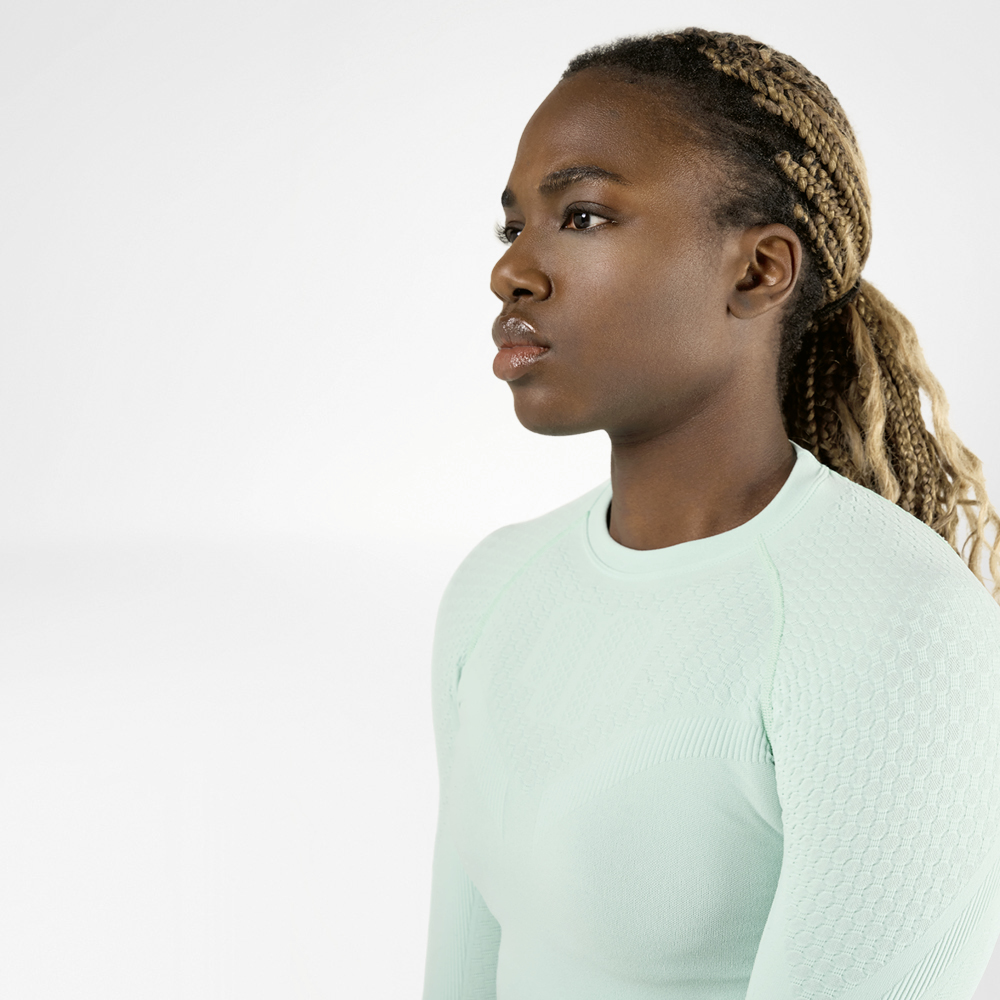Een sportieve vrouw met gevlochten haar die een lichtgroen, langmouwig compressieshirt draagt, wordt van opzij bekeken. Ze heeft een geconcentreerde en vastberaden uitdrukking op haar gezicht.