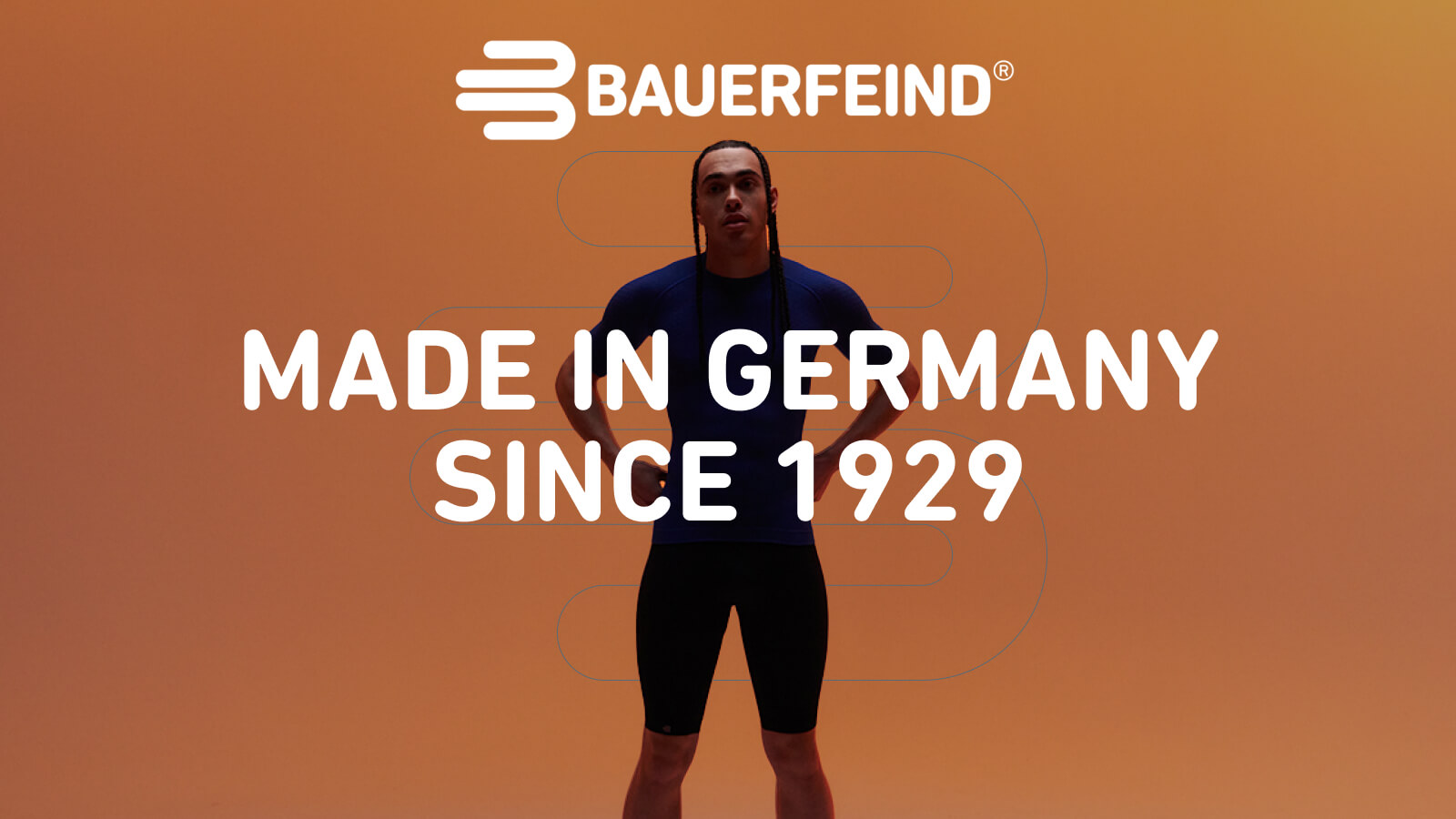 Zelfverzekerd in Bauerfeind compressiekleding Een persoon staat zelfverzekerd met iets gespreide benen voor een oranje achtergrond. Ze dragen een nauwsluitend blauw sportshirt en een zwarte sportbroek.