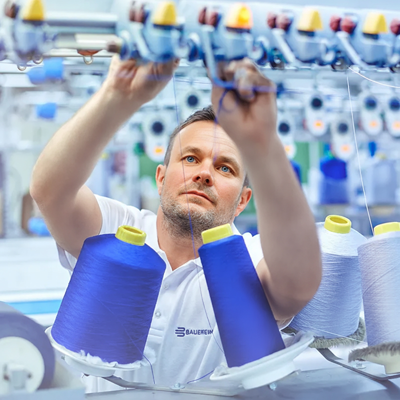 Een medewerker van Bauerfeind werkt geconcentreerd aan een breimachine en leidt blauwe garens door een nauwkeurig textielsysteem. Op de voorgrond zien we klossen garen in blauw, wit en geel – een teken van Duitse topkwaliteit.