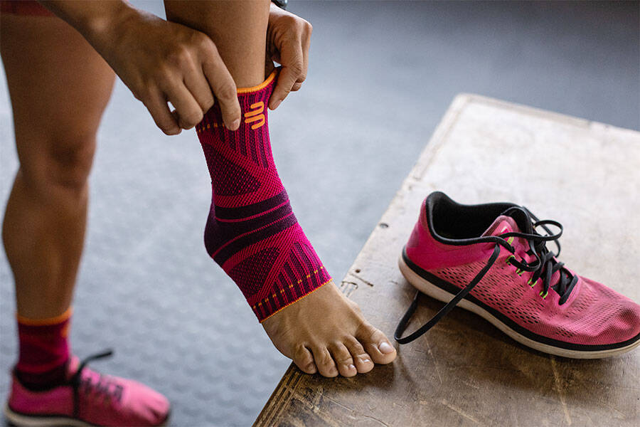 Met zijn flexibiliteit is Ankle Support Dynamic een geweldige metgezel tijdens de training Vrouw trekt een roze voettape aan en ondersteunt haar voet op een doos