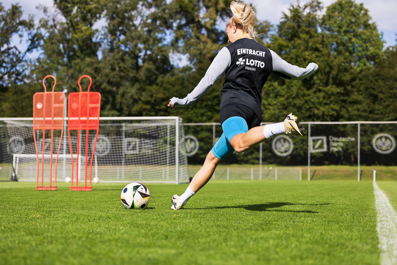 Speelster van Eintracht Frankfurt tijdens schietoefening Een speelster van Eintracht Frankfurt schiet met volle kracht op het doel op de achtergrond. Ze draagt trainingskleding met het Eintracht-logo en een blauwe knieband van Bauerfeind Sports.