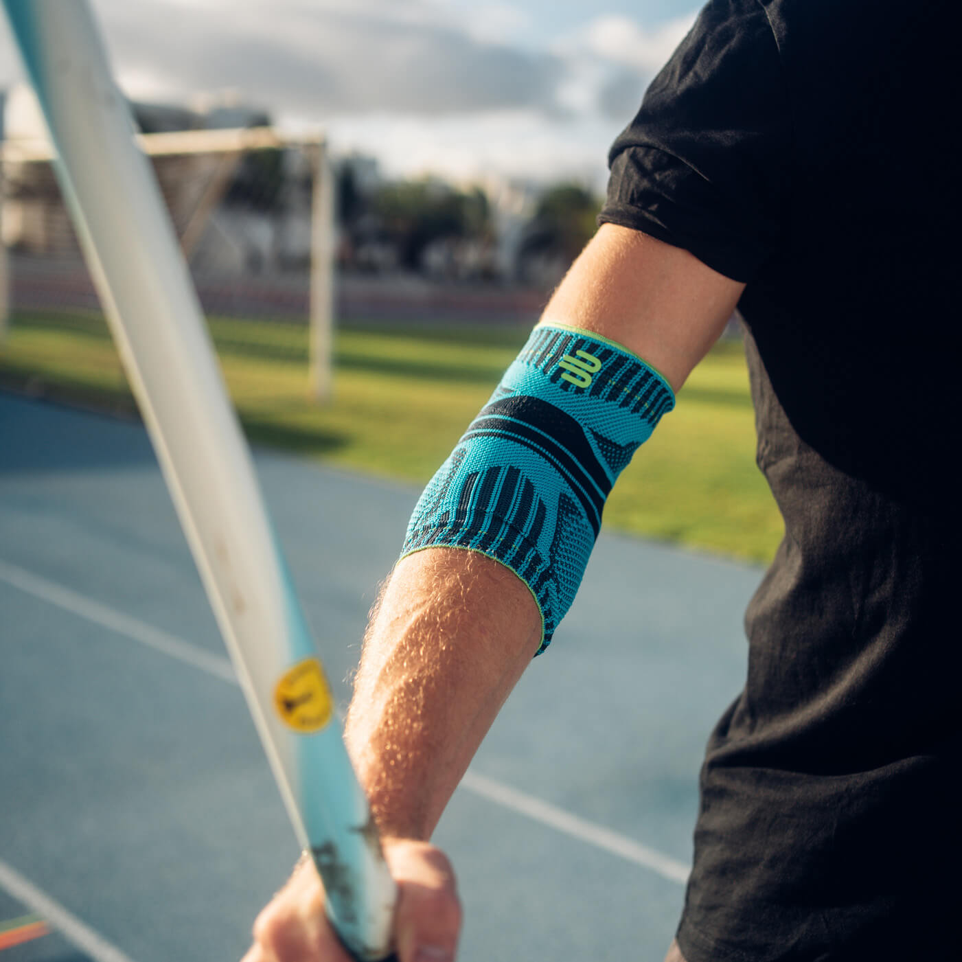 Precisie en kracht – met stabiele ondersteuning voor je elleboog. Close-up van een arm met een blauwe elleboogbrace tijdens het speerwerpen. Op de achtergrond zijn een atletiekbaan en een zonnig landschap te zien.